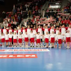 Grupa młodych ludzi w strojach piłki ręcznej stoi przed dorosłymi zawodnikami. Wszyscy ubrani są w białe koszulki i czerwone spodenki.