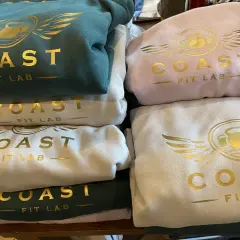 Stosik bluz klubowych w kolorach zielonym, różowym i kremowym, z złotym logo "COAST FIT LAB".