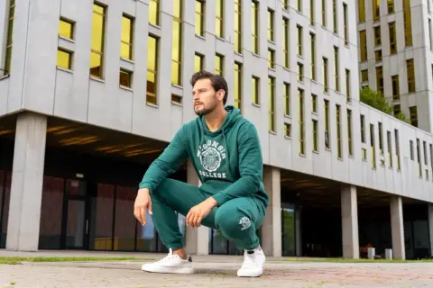 Mężczyzna w zielonym dresie Napoli siedzi na ziemi przed nowoczesnym budynkiem.