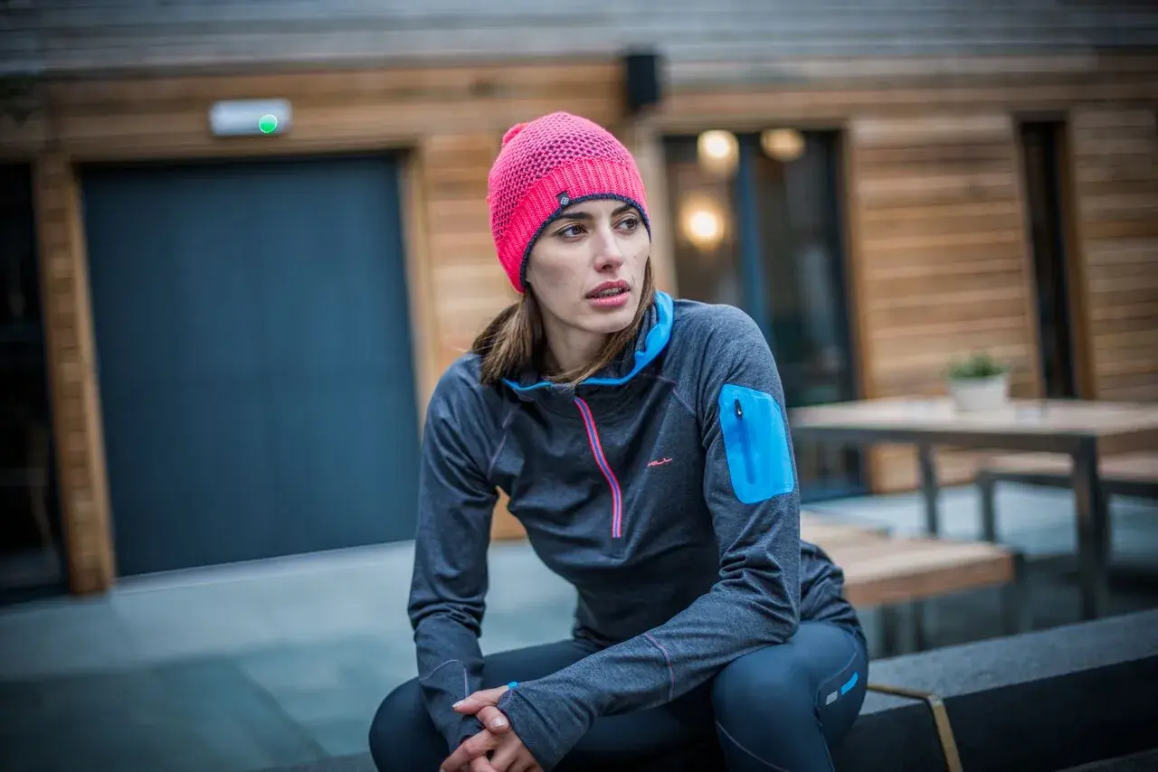 Kobieta w sportowym stroju, z jaskrawym, różowym czapka beanie, siedzi na ławce przed nowoczesnym budynkiem.