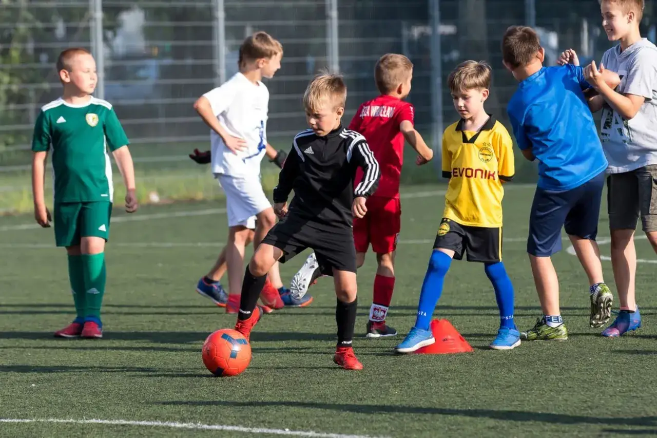 Młodzik wiek na boisku piłkarskim, ćwiczący drybling. Dzieci w kolorowych strojach sportowych, skupione na grze.