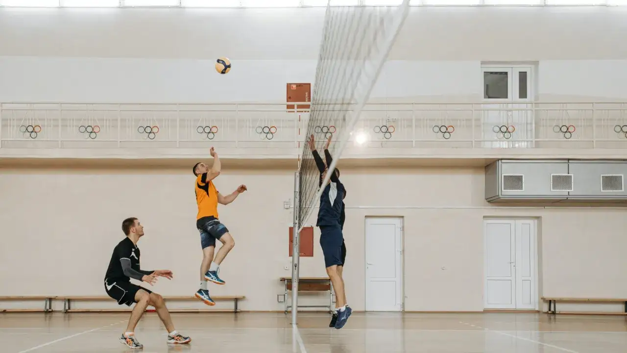 Trzech siatkarzy w akcji. Jeden z nich skacze do zbicia, drugi serwuje, a trzeci przygotowuje się do obrony. W tle widnieją olimpijskie kółka. Ile jest setów w siatkówce? Zależy od wyniku!